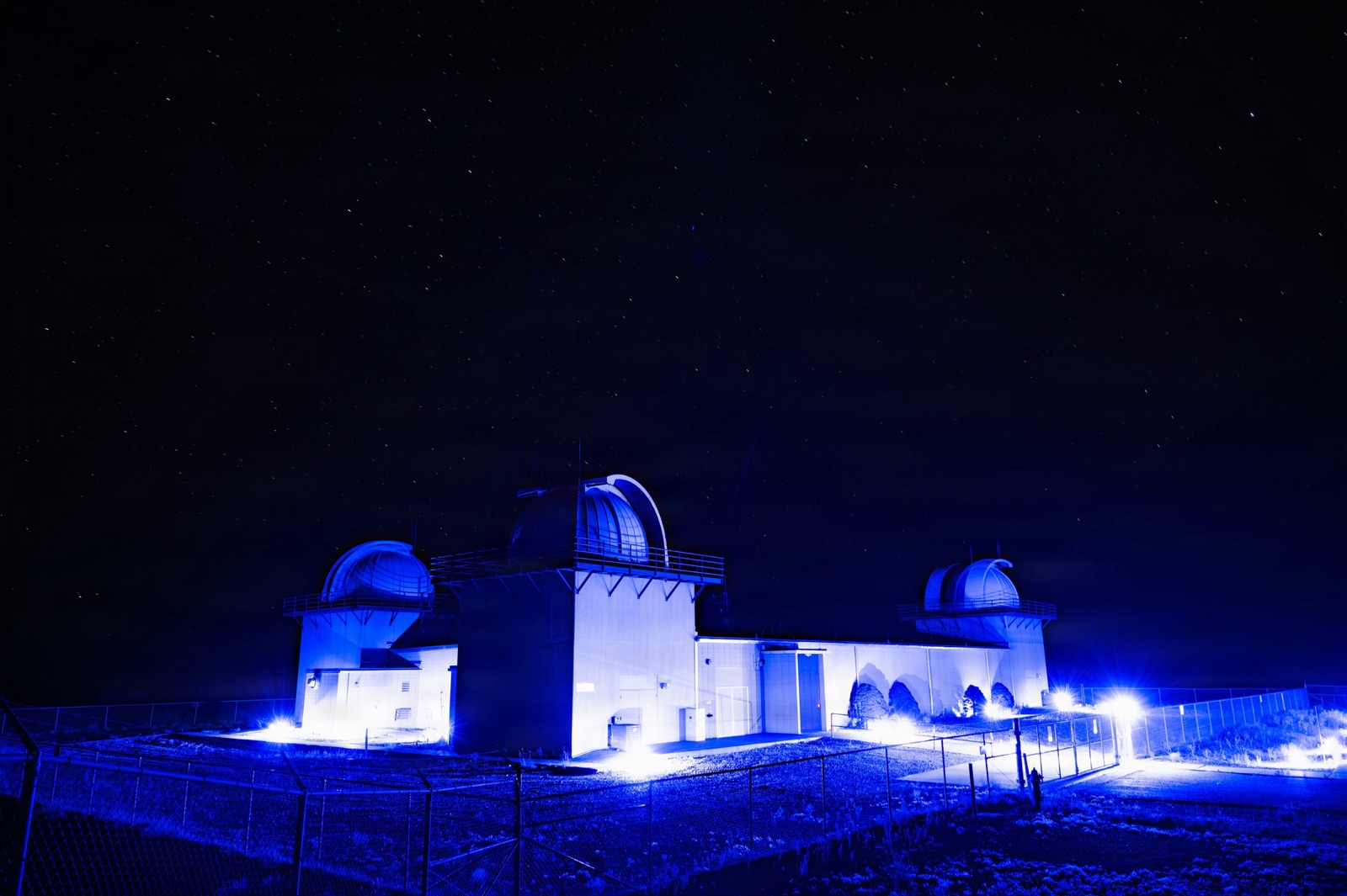 Three Ground-based Electro-Optical Deep Space Surveillance telescope domes at White Sands, New Mexico, lit blue at night under a starry sky.